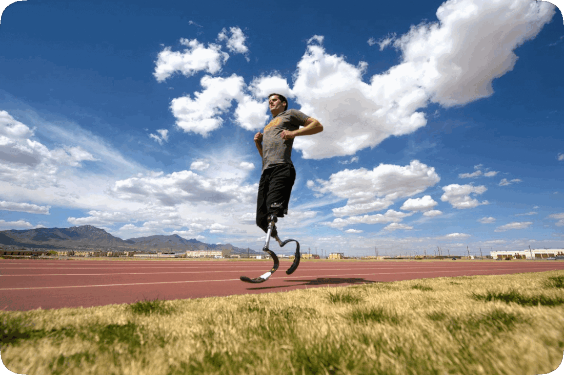 Picture of a double-amputee wearing prosthetics and running on a track that is bordered on both sides by green grass, with hills in the distance and a blue sky and fluffy white clouds overhead.