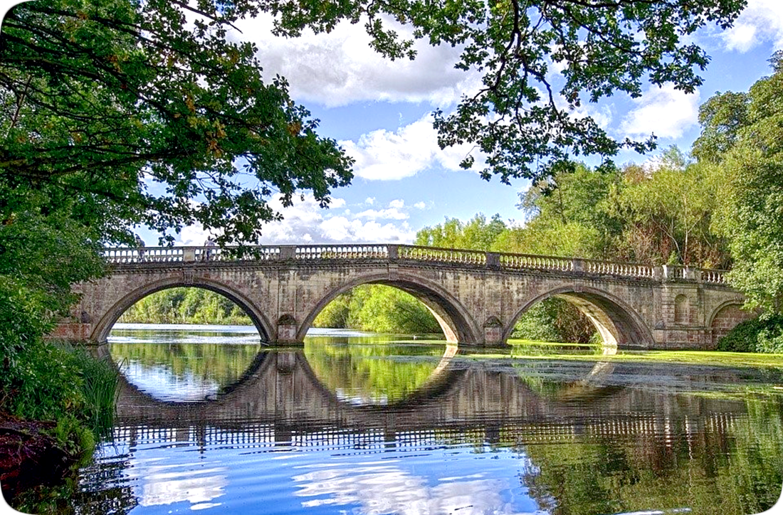Bridge over calm waters, surrounded by trees with green leaves and blue sky with fluffy white clouds.